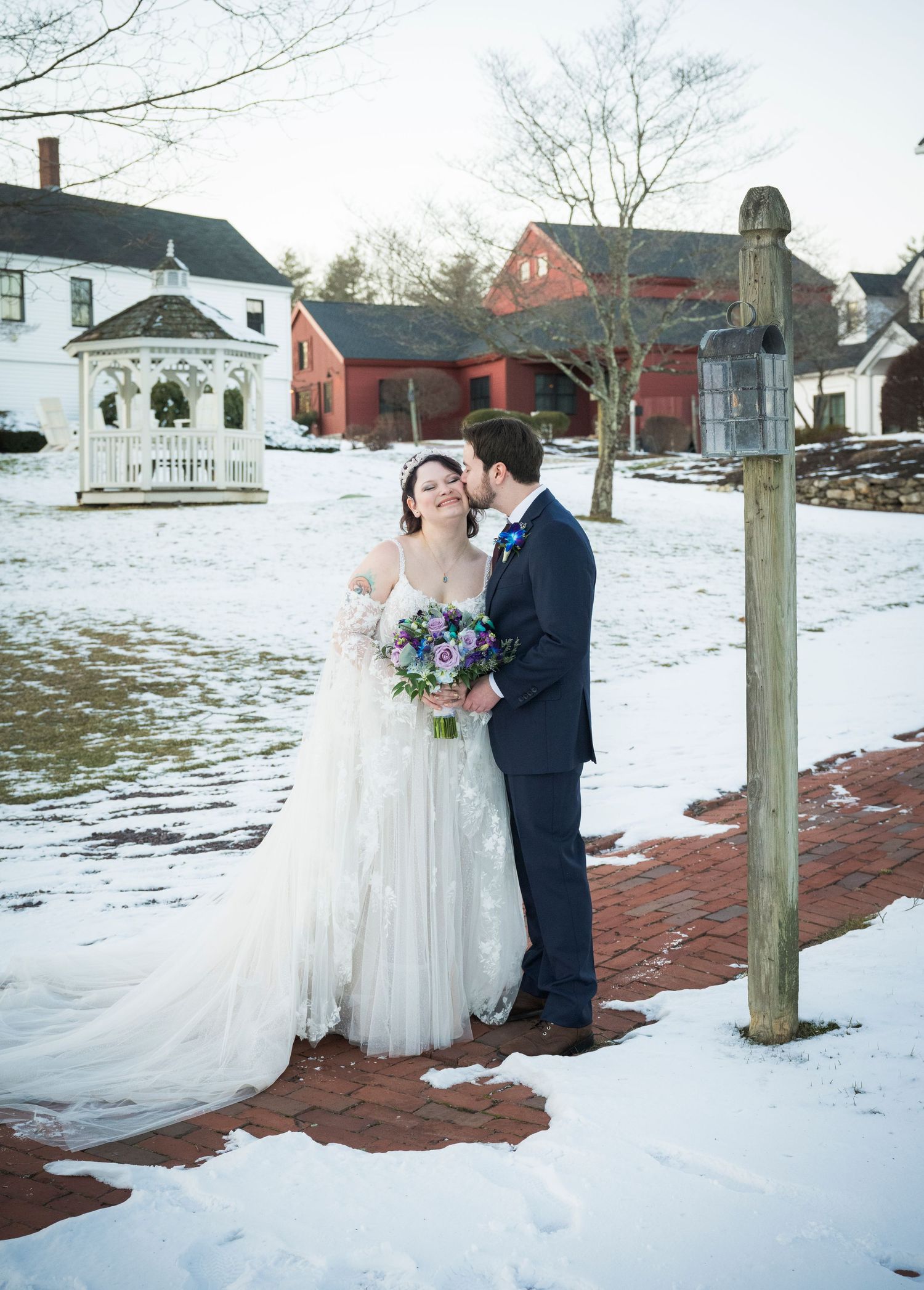 A couple shares a romantic winter wedding moment on a snow-covered brick path beside historic buildings.