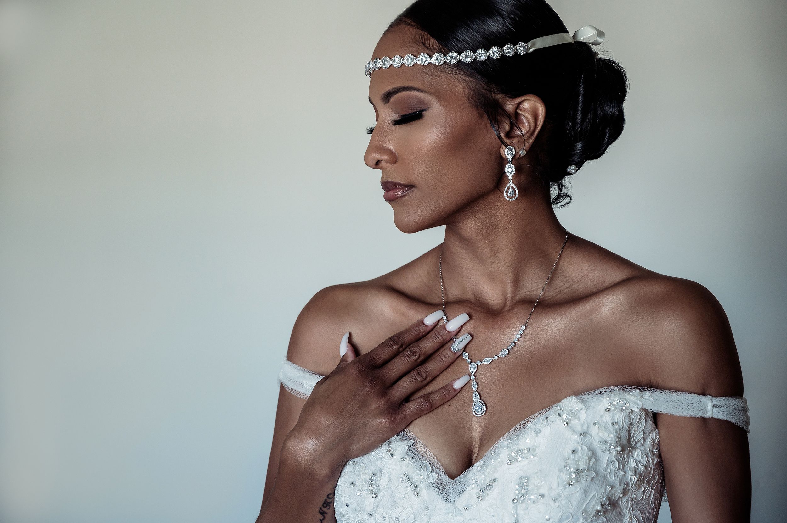 Elegant bride in off-shoulder white gown wearing crystal headpiece and jewelry with eyes closed in profile view.