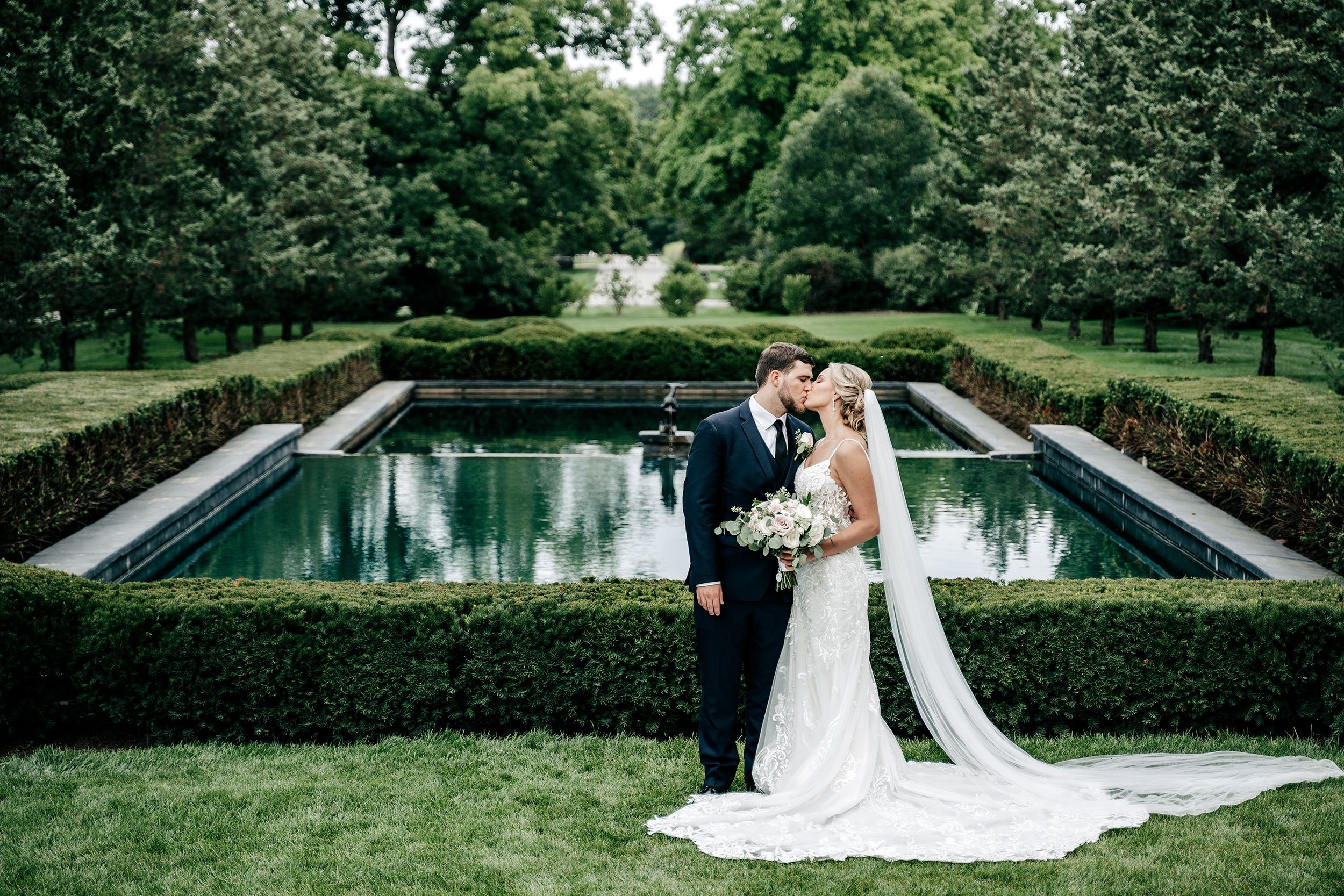 A couple poses in a formal garden setting with manicured hedges and reflecting pools during a wedding celebration.