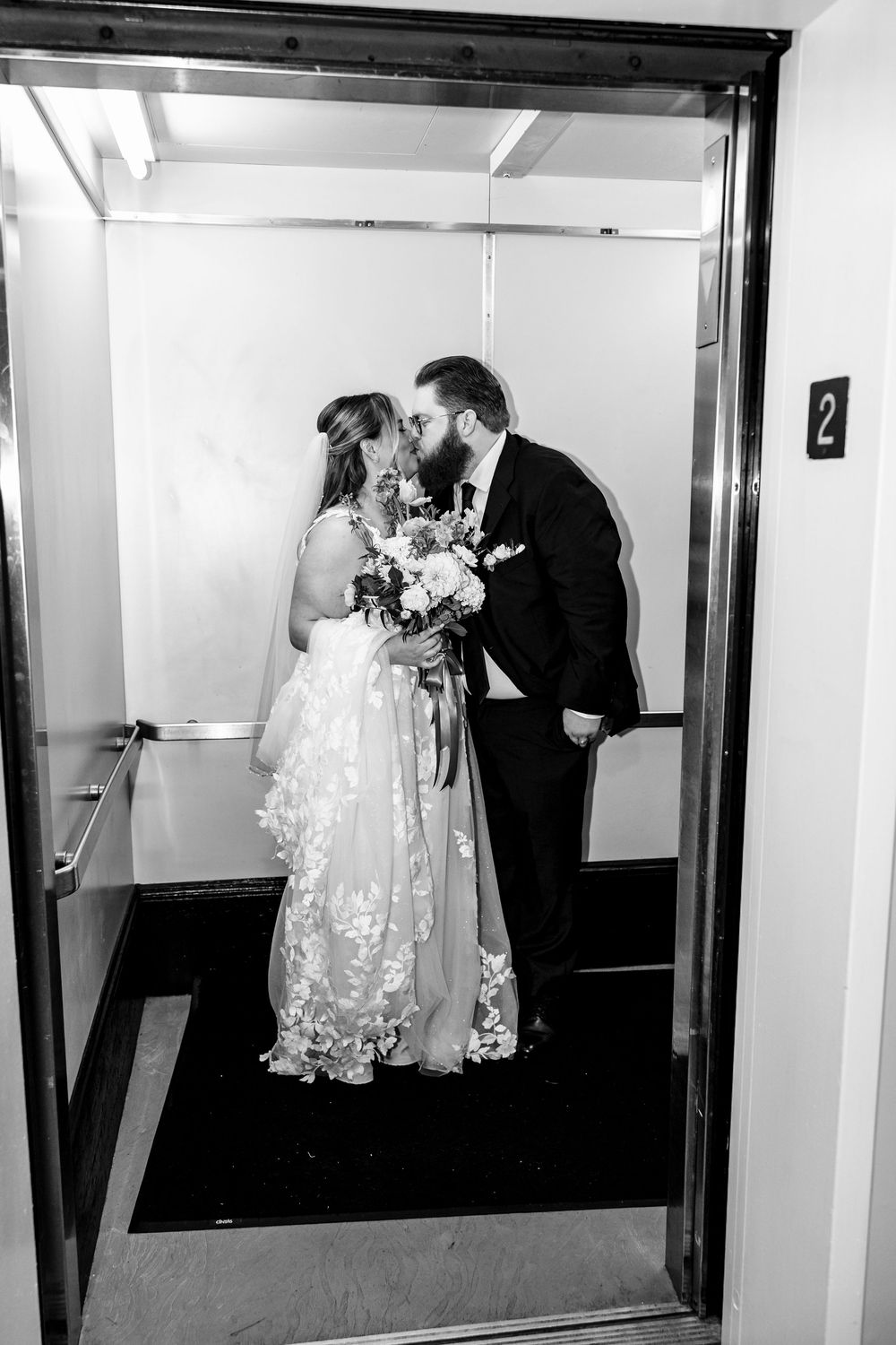 A romantic black and white photo of newlyweds sharing a kiss in an elevator while the bride holds a floral bouquet.