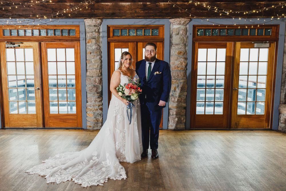 A newlywed couple poses in front of rustic windows with string lights, the bride wearing a lace gown with a long train.