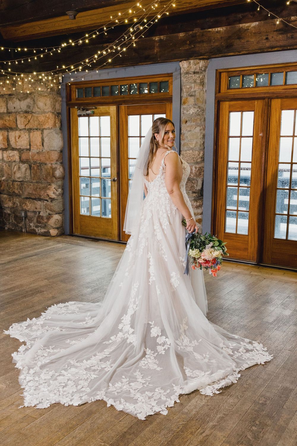 Stunning bridal gown with intricate lace details and dramatic cathedral train displayed in rustic venue with string lights.