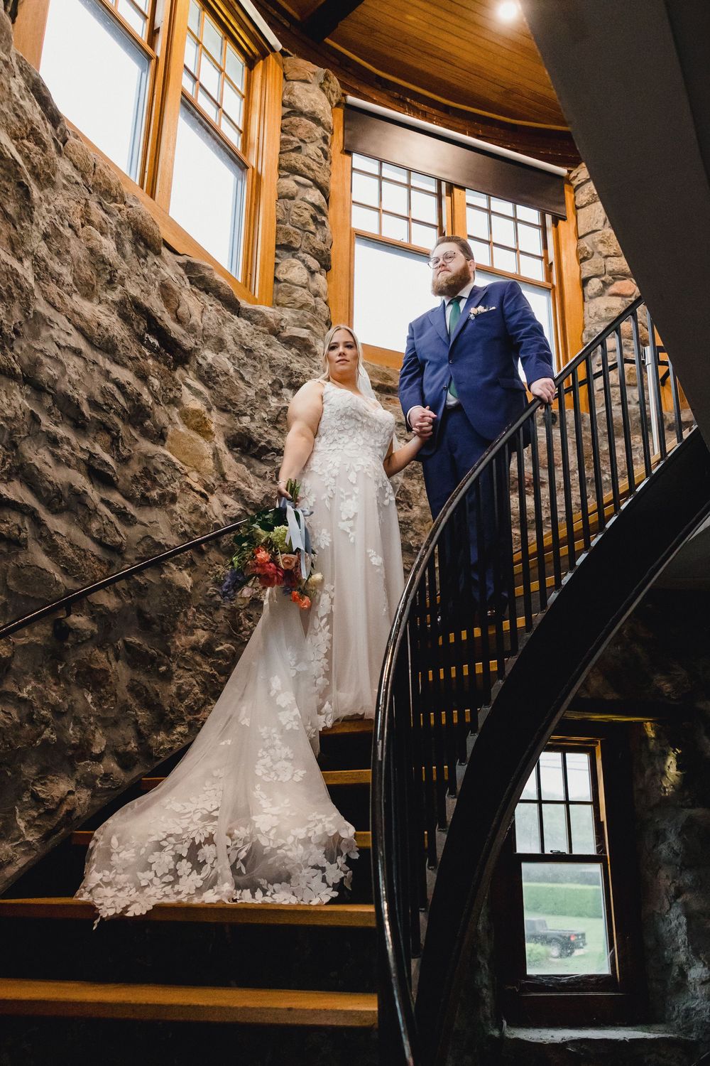 A couple poses on an elegant wooden staircase with stone walls in a rustic indoor setting.