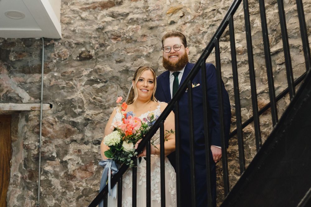 A wedding couple poses on stone steps with a rustic stone wall backdrop and warm lighting.