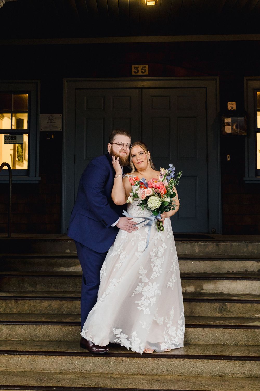 A couple in wedding attire share an intimate moment while sitting on stone steps at dusk.