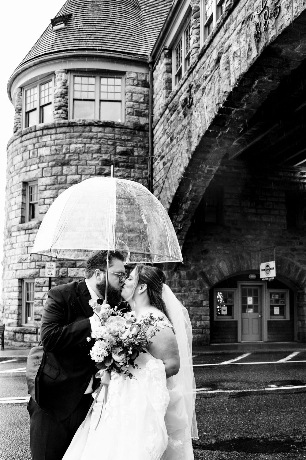 A couple shares a romantic moment under a clear umbrella against a stone building backdrop.