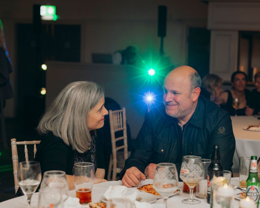 Two people enjoy conversation while dining at an evening event with colorful ambient lighting and table settings.