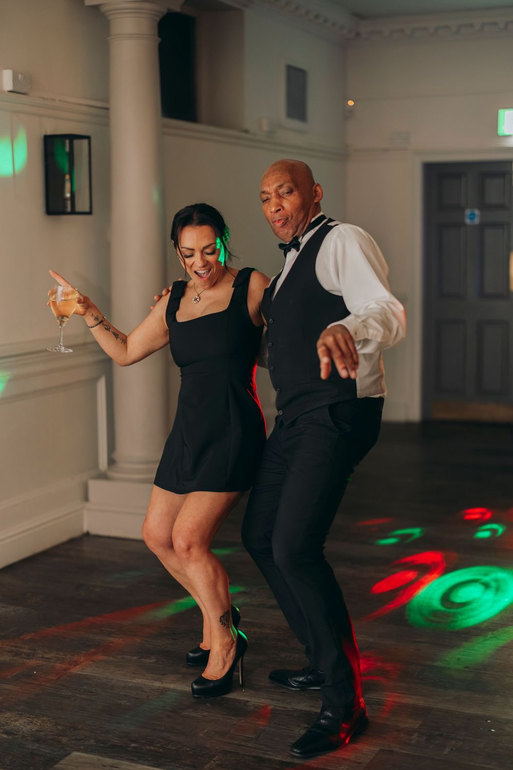 Two dancers perform energetic swing dance moves on a wooden floor with colorful lights reflecting in the background.
