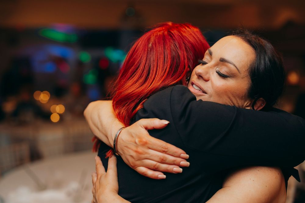 Two people share an emotional embrace at a dimly lit event with colorful lights in the background.