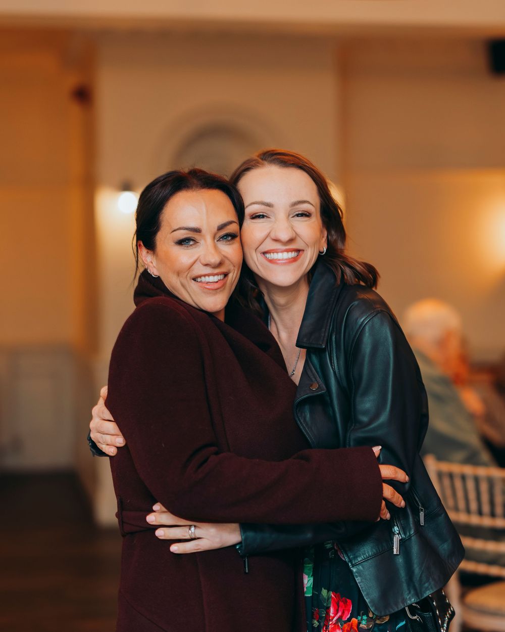 Two people embrace warmly in an indoor setting with warm lighting and a joyful atmosphere.