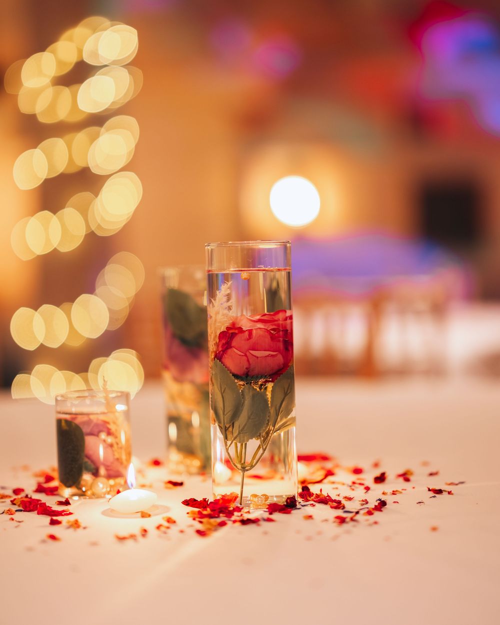 Red rose petals scattered on a table with glowing candlelight and bokeh effects create a romantic atmosphere.