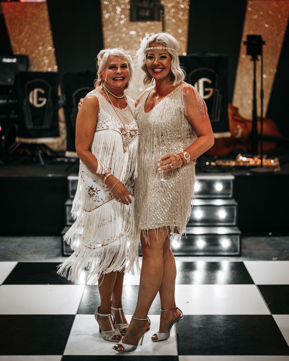 Two people in sparkly white flapper-style dresses dancing together on a black and white checkered floor.
