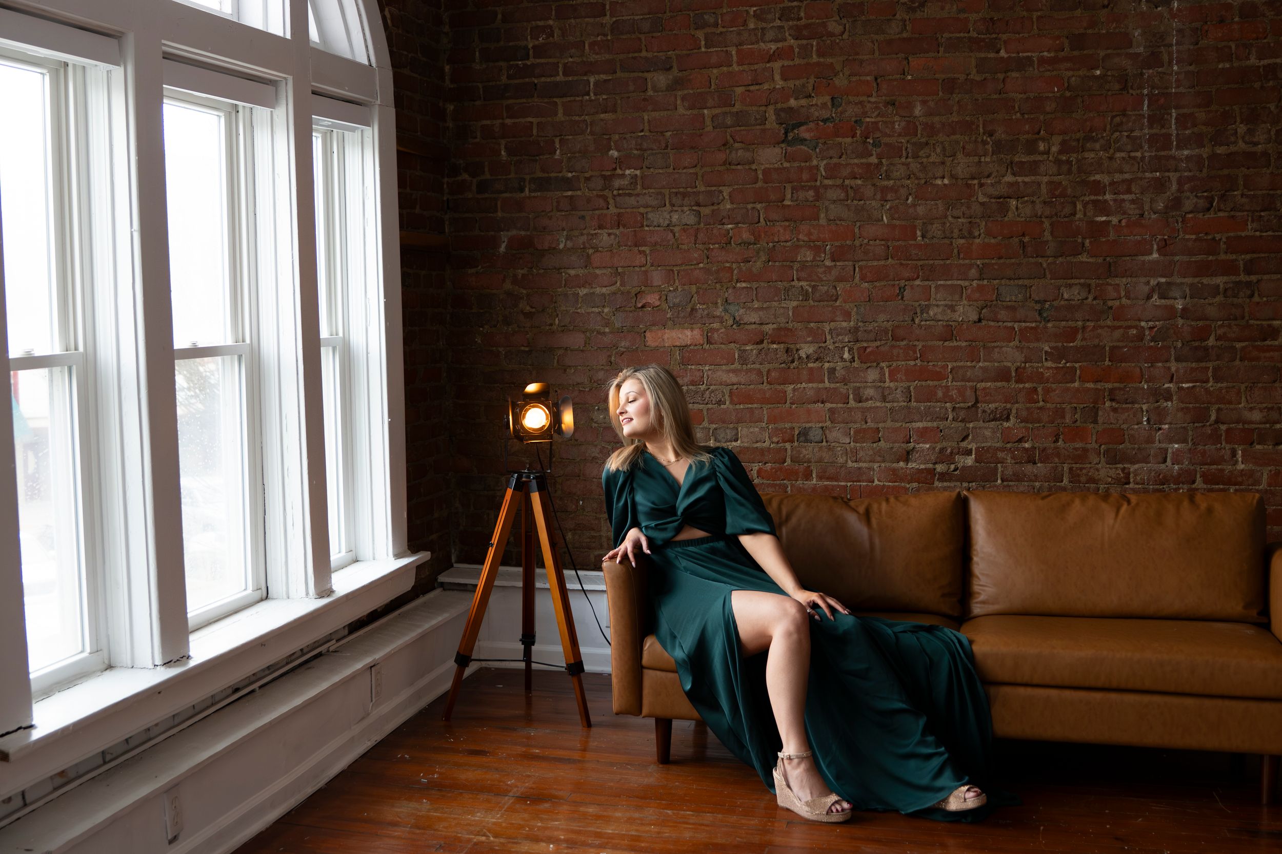 A person in a dark dress sits by a window in a rustic brick room with natural lighting.