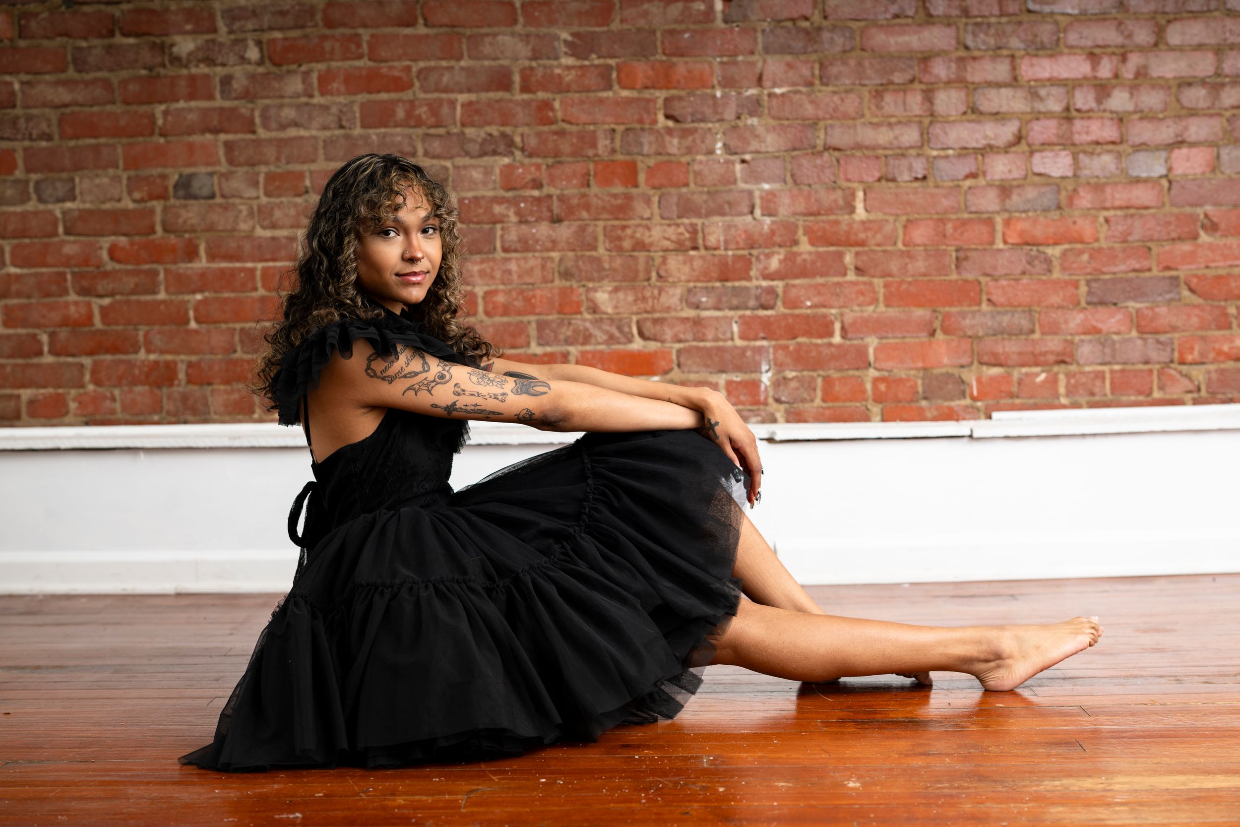 A dancer in a black dress performs a graceful floor sequence against a rustic brick wall backdrop.