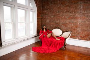 A figure in a flowing red dress poses gracefully against exposed brick walls in a sunlit studio space.