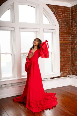A person in a flowing red dress poses dramatically against white arched windows and exposed brick walls in a studio.