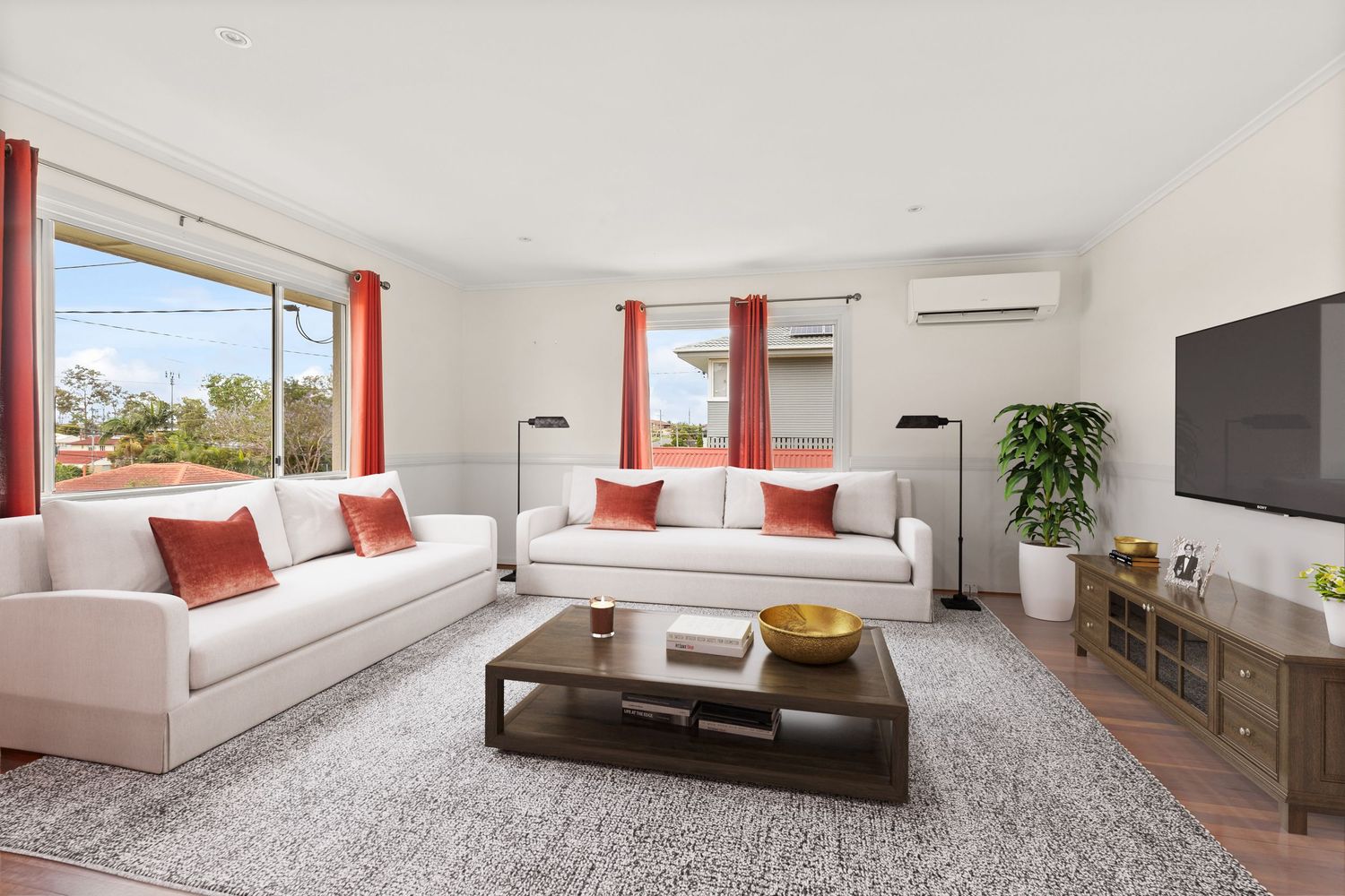Bright living room with white sofas, red cushions, split-system AC, timber floors, and large windows.