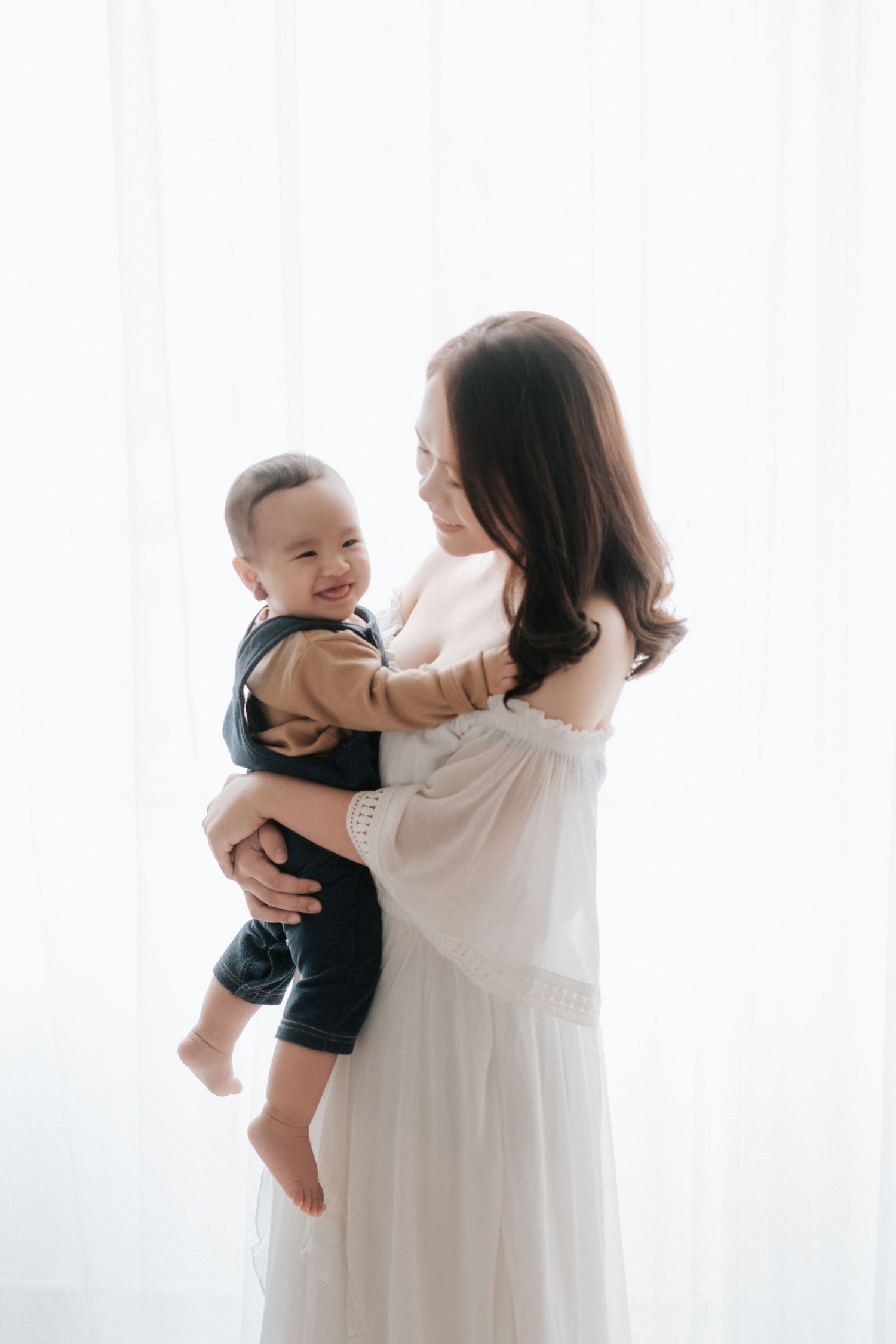 A smiling mother in a white dress holds her happy baby boy in a bright, softly lit studio setting.在明亮柔和的燈光下，一位身穿白色連身裙、微笑的母親抱著她快樂的男嬰。