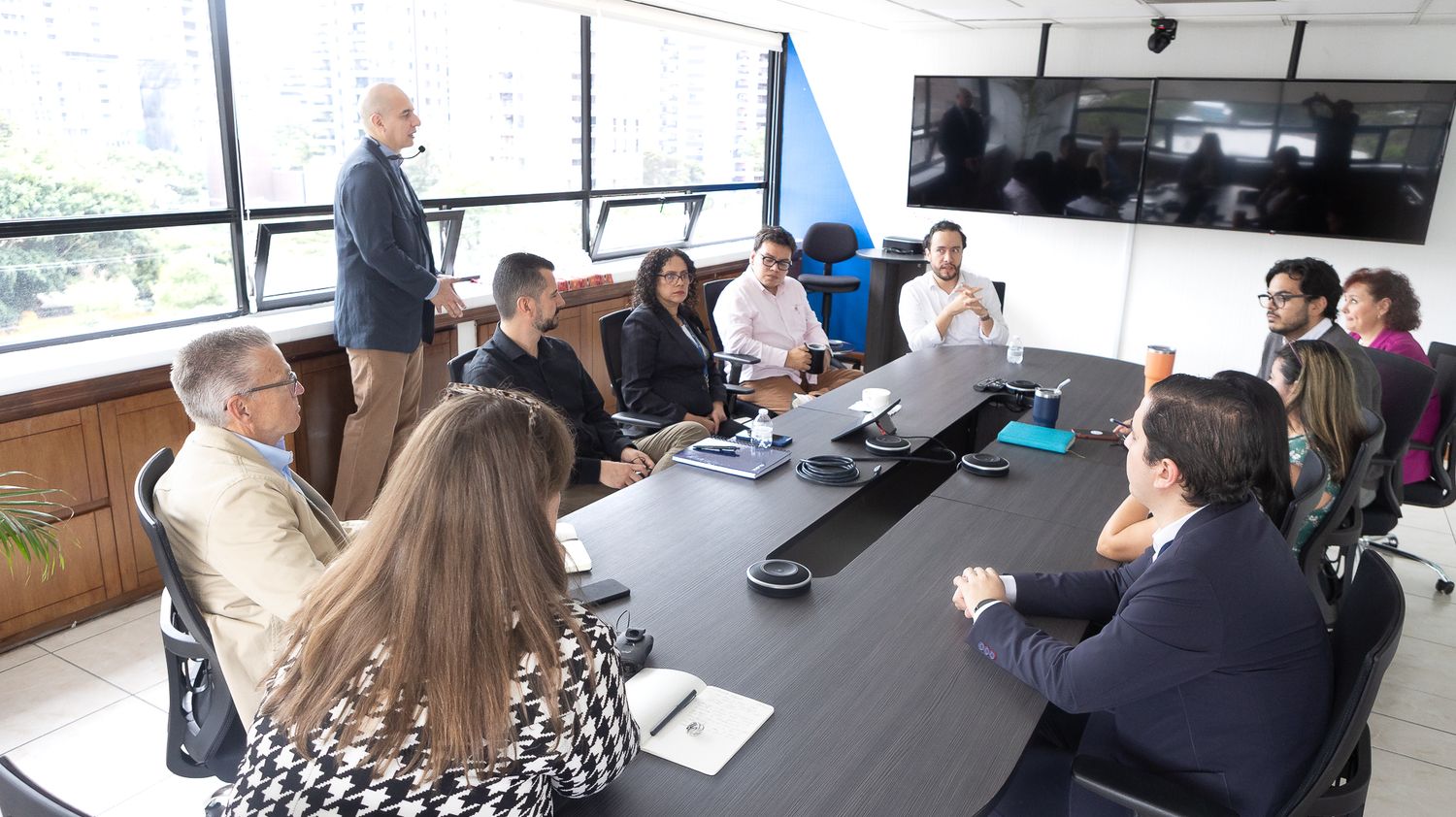 People gathered around a conference table in a modern meeting room with large windows and presentation displays.