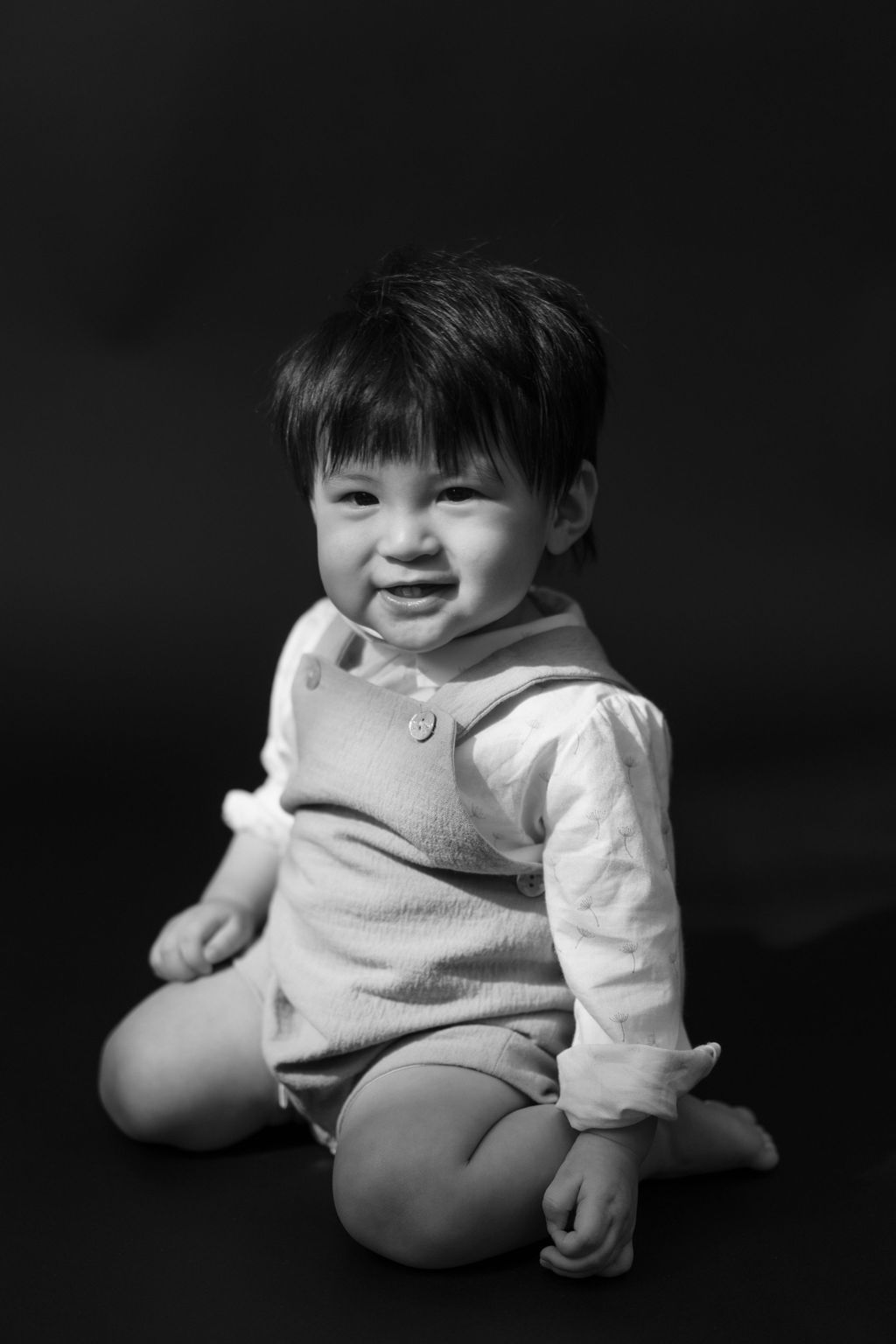 Black and white portrait of a smiling toddler sitting cross-legged wearing overalls and a long-sleeve shirt一張黑白肖像照，照片中一個微笑的幼兒盤腿坐著，穿著背帶褲和長袖襯衫