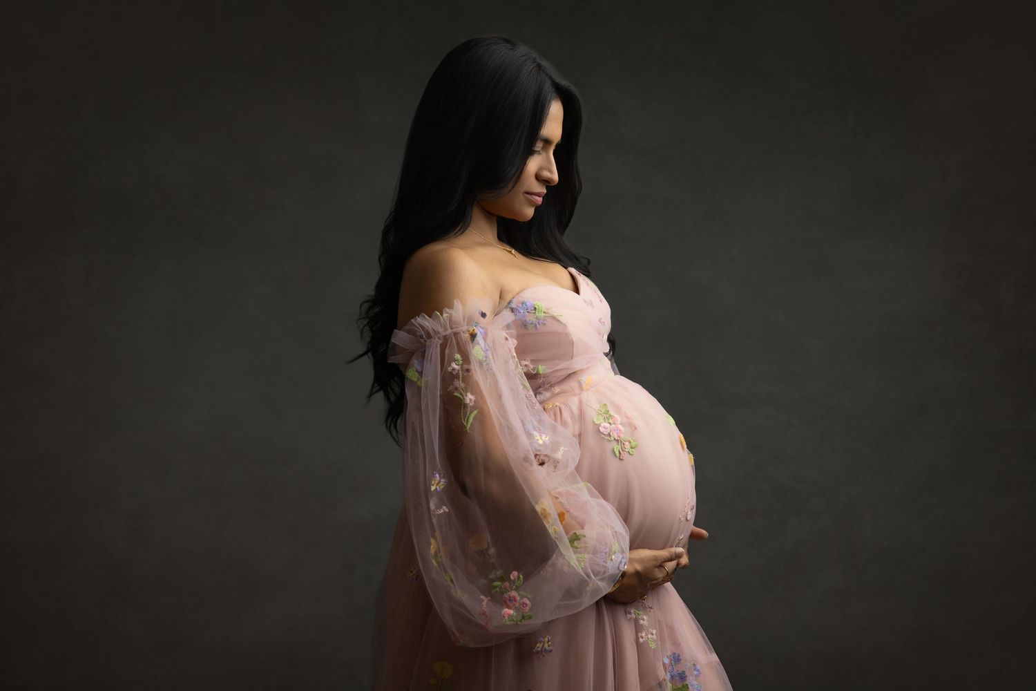 A woman in a romantic pink tulle gown poses against a dark backdrop in a maternity photoshoot.