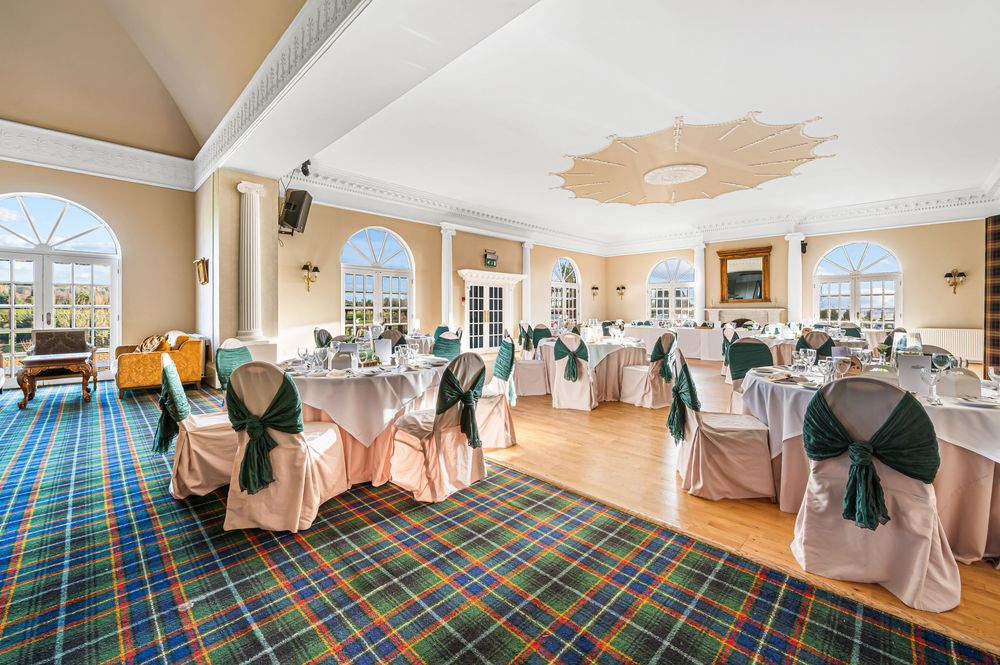 Elegant banquet hall setup with white chair covers, teal sashes, and plaid carpeting in a bright sunlit room.