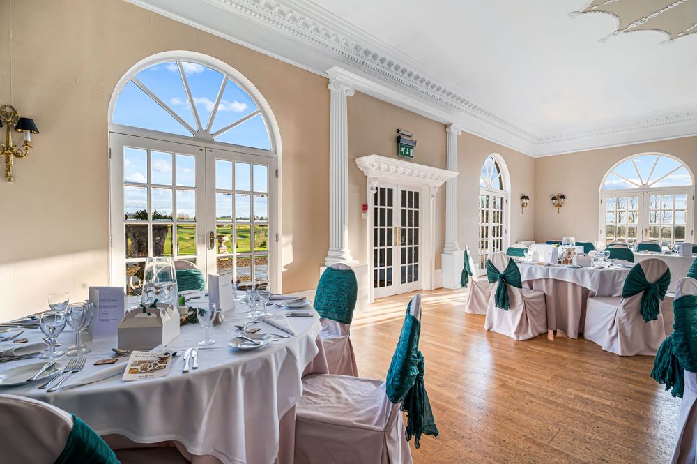 Elegant banquet hall with large arched windows, white tablecloths, and teal chair sashes in a bright sunlit room.