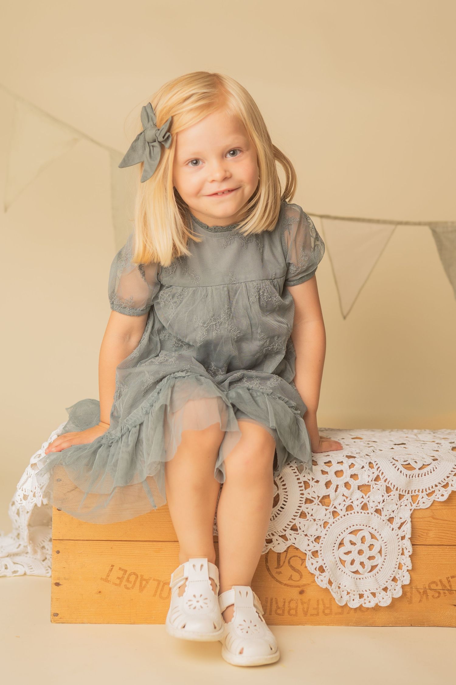 A child in a gray dress and bow sits on a wooden crate with lace doilies in a vintage-style portrait setting.