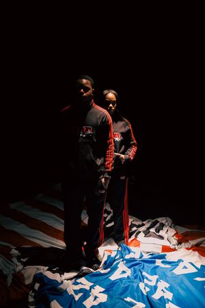 Two figures in black and red tracksuits stand dramatically on a surface covered with blue and white fabric in dark lighting.