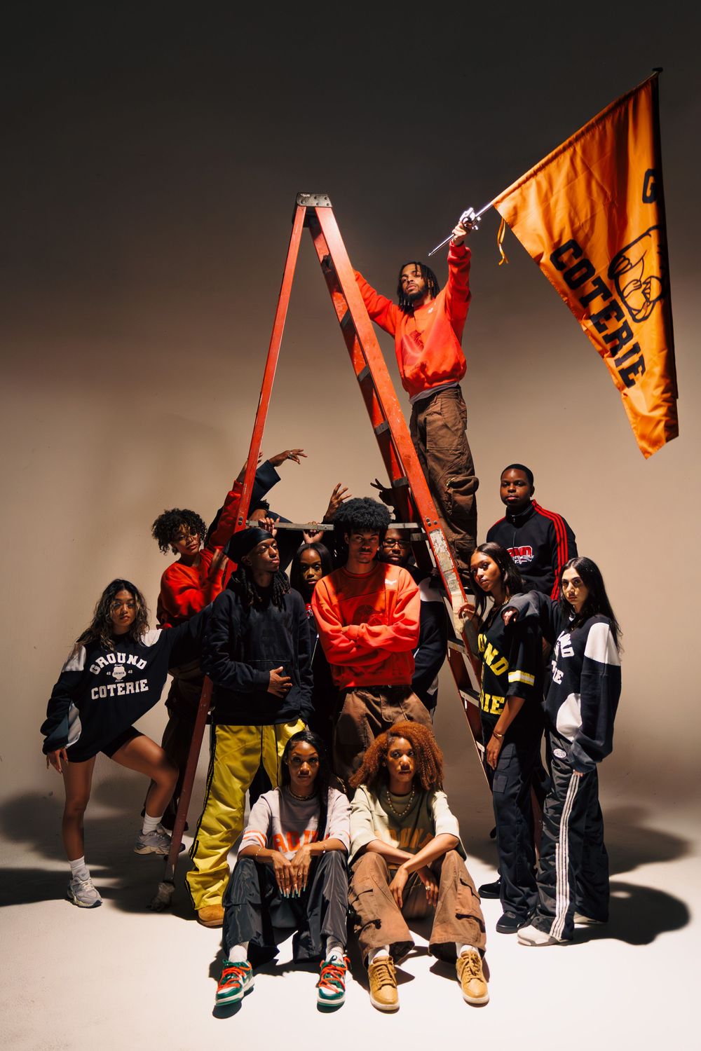 Group photo of people in urban streetwear posing with a ladder and orange flag against a studio backdrop.