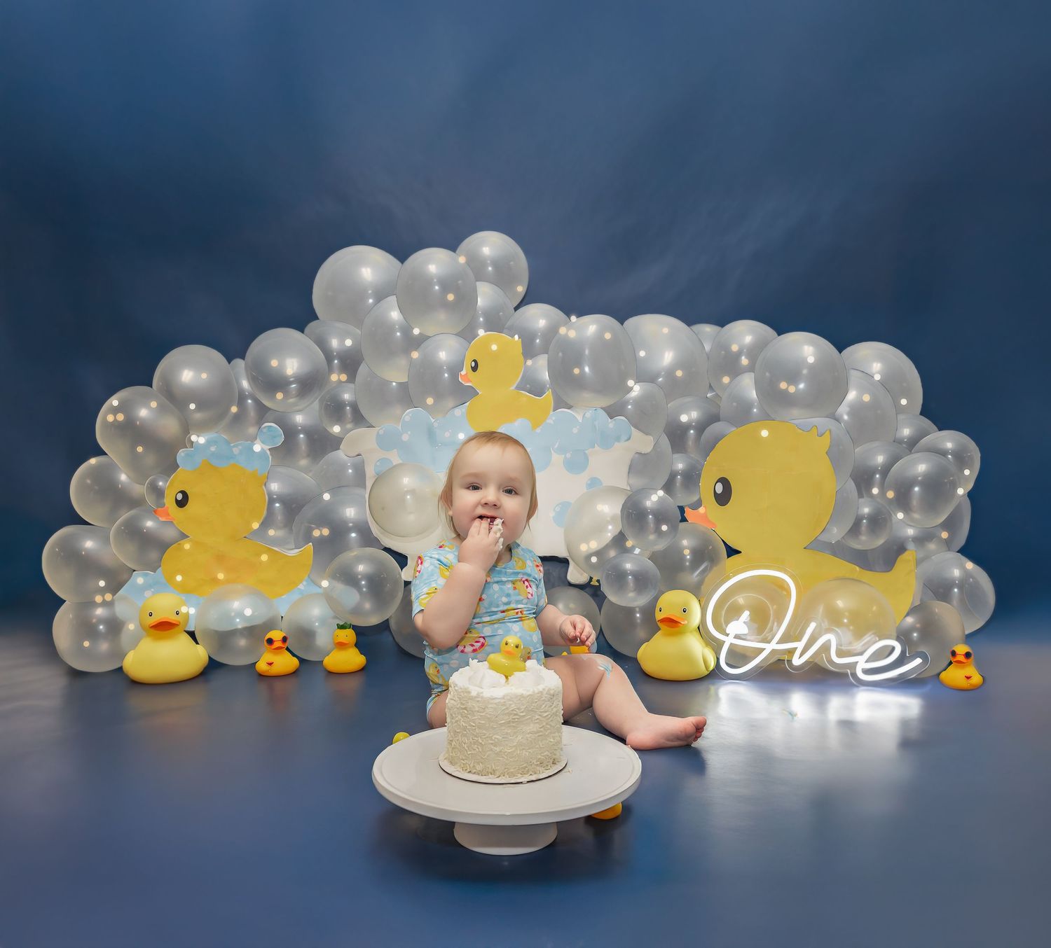 Rubber duck themed cake smash setup with silver balloon arch and white cake.