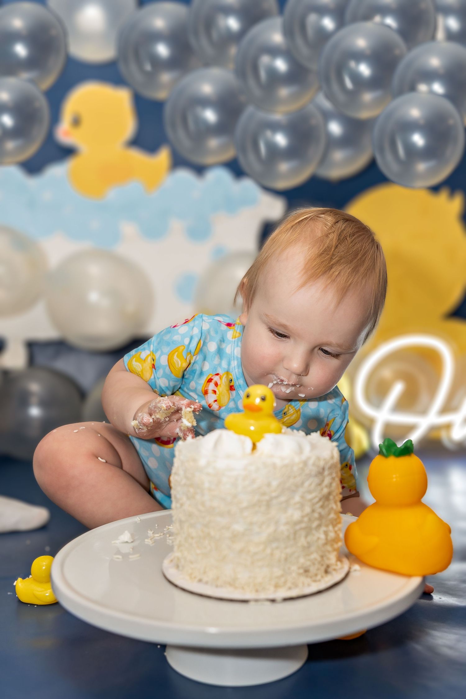 White birthday cake with rubber ducky decorations and silver balloon backdrop.