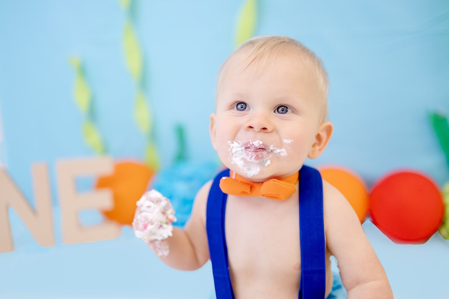A baby in a blue outfit celebrates their first birthday with colorful cake smash photos.