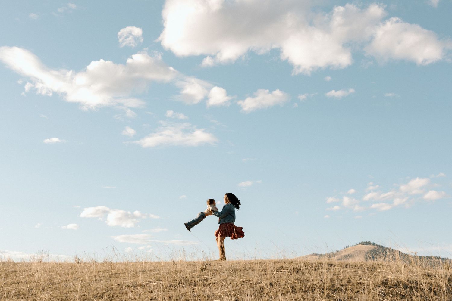 Figure leaping with arms raised against blue sky and white clouds on a hilltop.