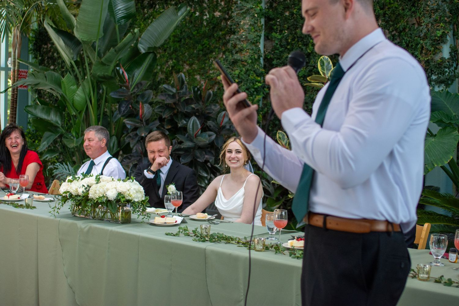 Wedding party seated at head table with greenery garland decor while guest takes photo on phone during reception.