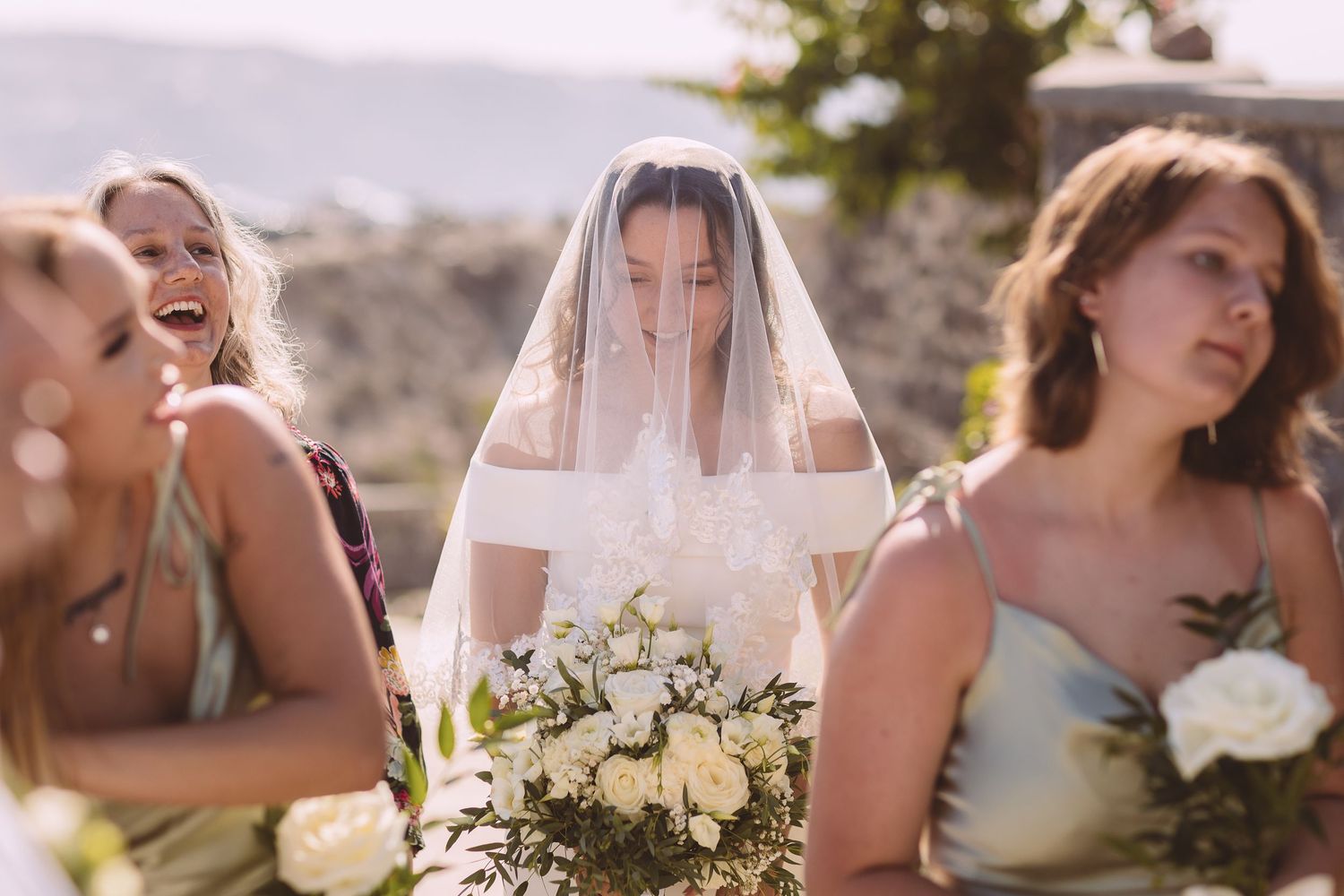 A Santorini bride with a veil viewed between bridesmaids looks excited before her destination wedding.