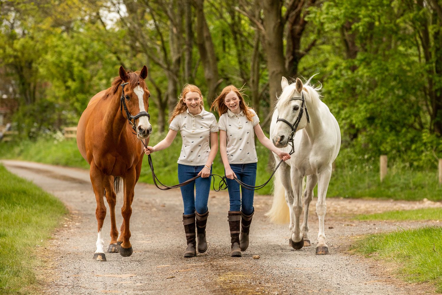 Three children walk with brown and white horses down a tree-lined path on a sunny day.