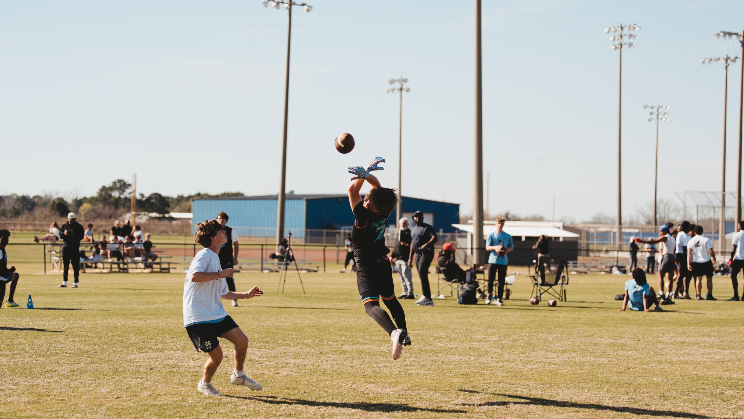 Athletes playing football on a sunny field with stadium lights in the background.