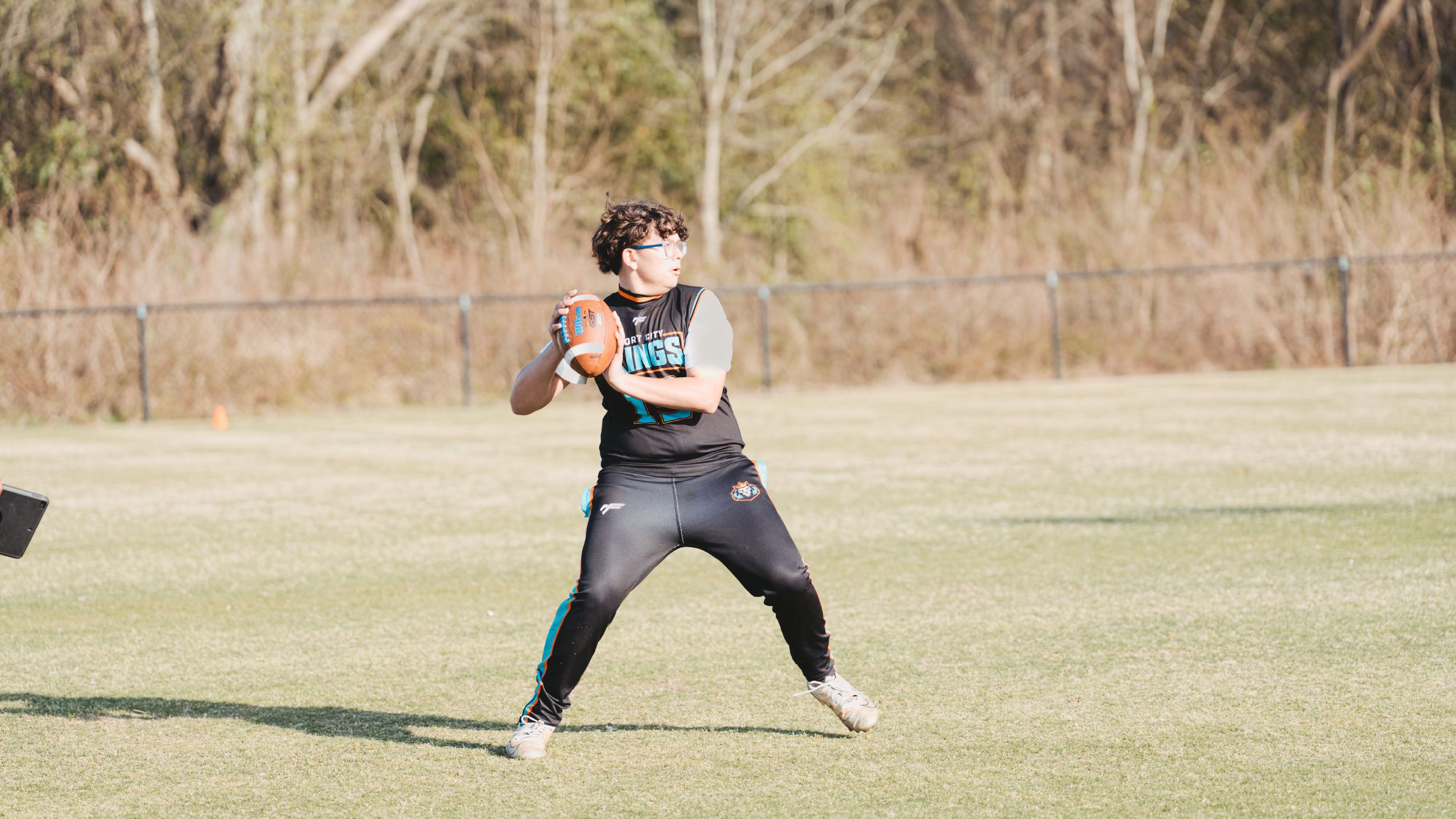 Athletes playing ultimate frisbee on a grassy field with trees in the background.