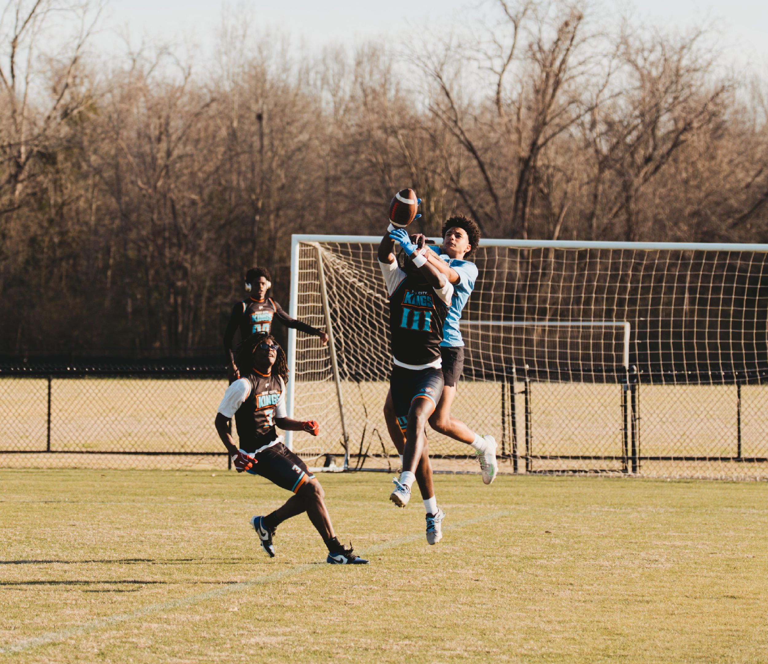 Soccer players compete for the ball during a match on a sunny day with goal posts in the background.