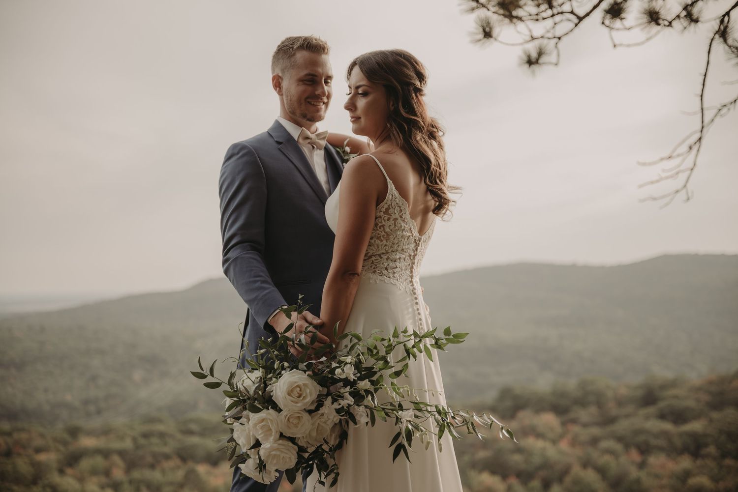 Elegant wedding couple poses with white floral bouquet against misty mountain landscape.
