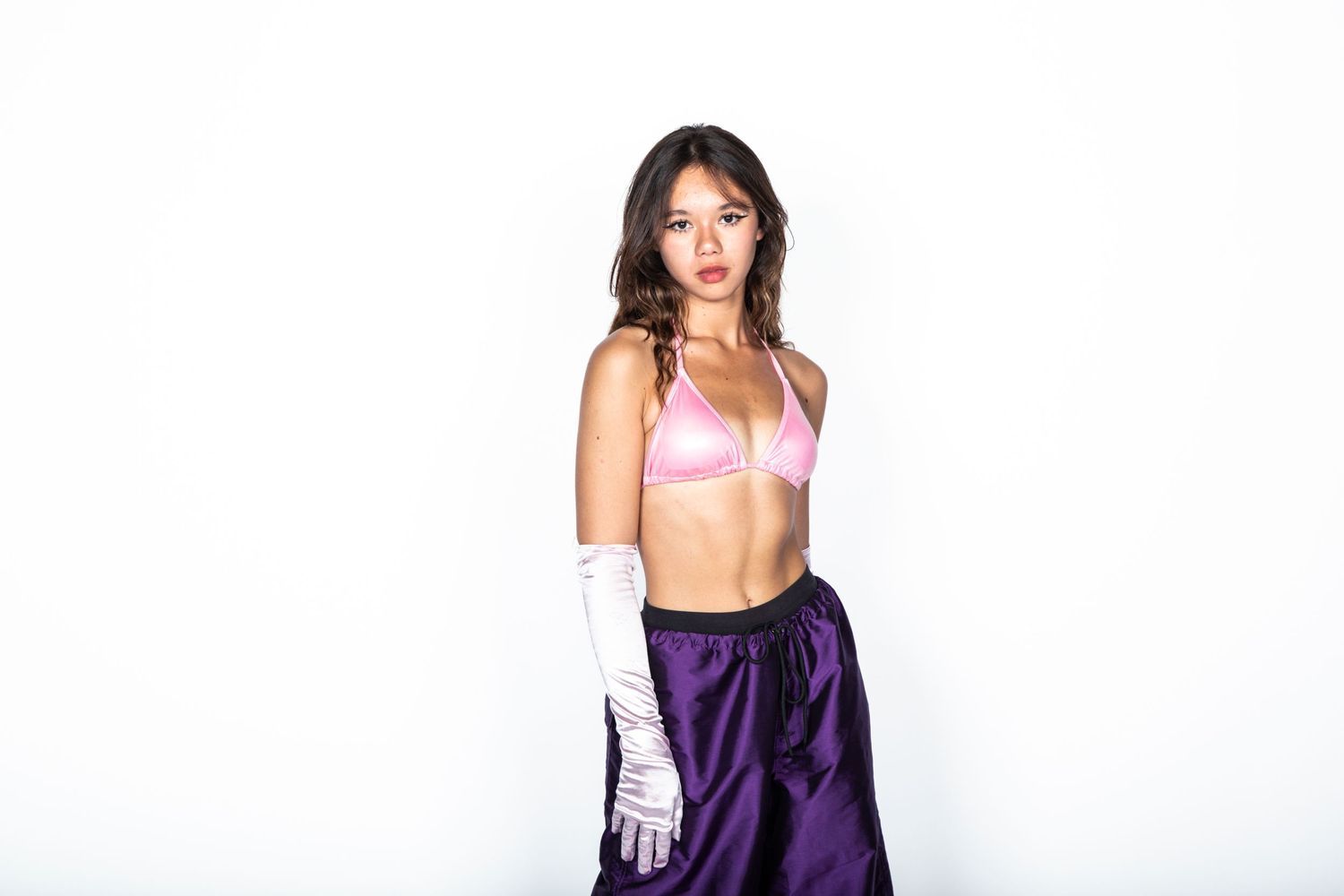 Fashion model wearing pink sports bra and purple satin skirt with white gloves against white background.