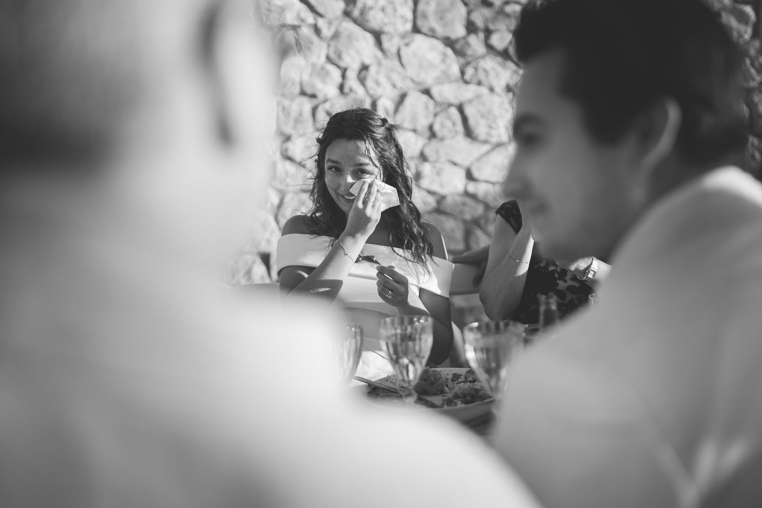 A bride dabs tears with a napkin between two guests at a Santorini wedding.