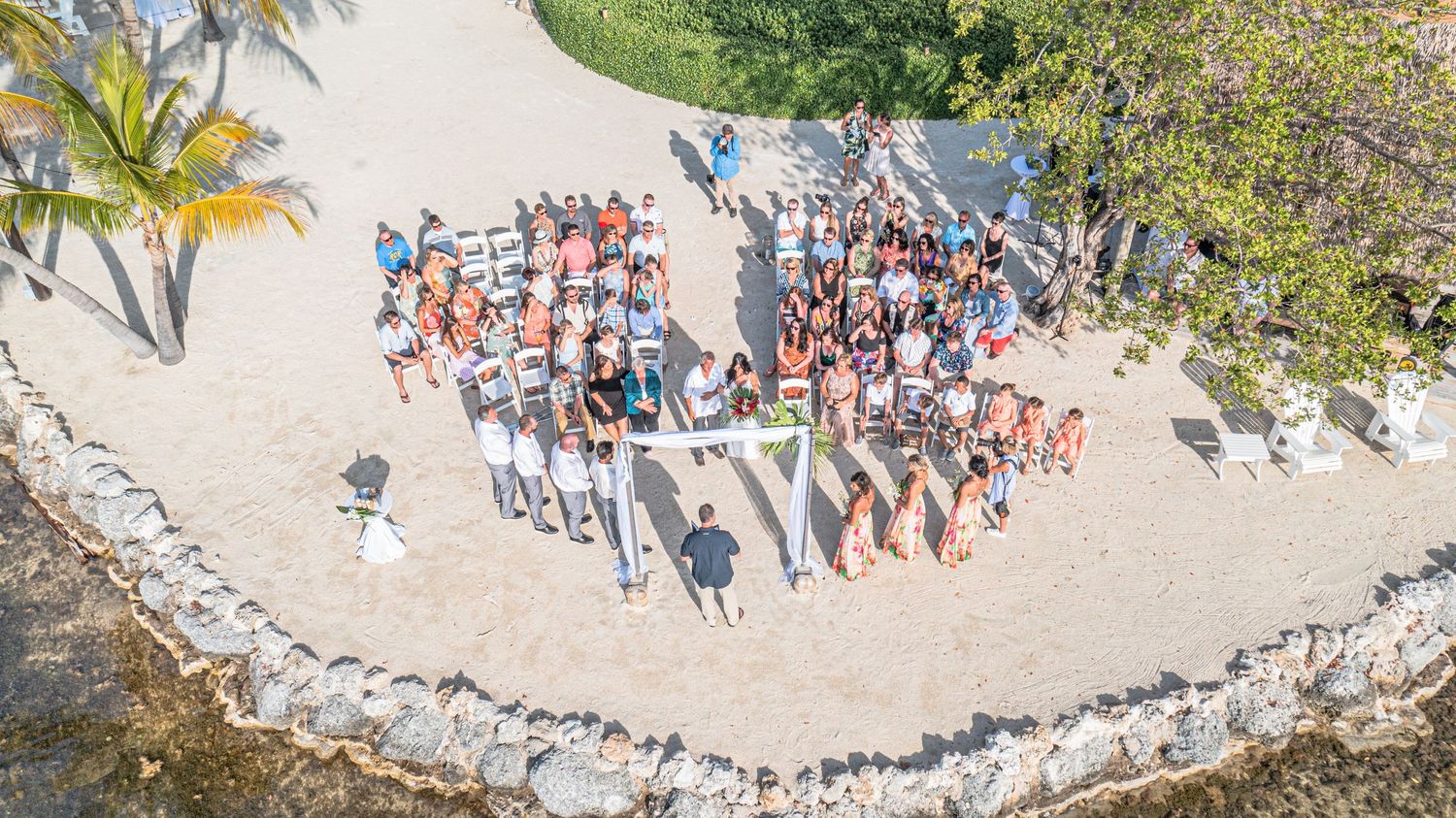 A scenic aerial drone shot reveals a large crowd of wedding guests gathered around an ocean bay on a tropical beach.