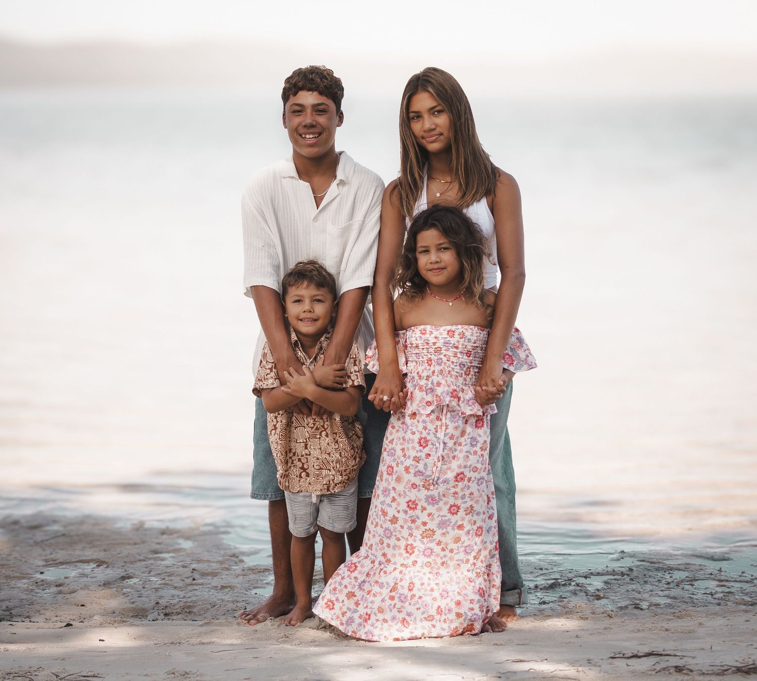 A family of four poses together on a beach, parents standing behind two young children smiling.