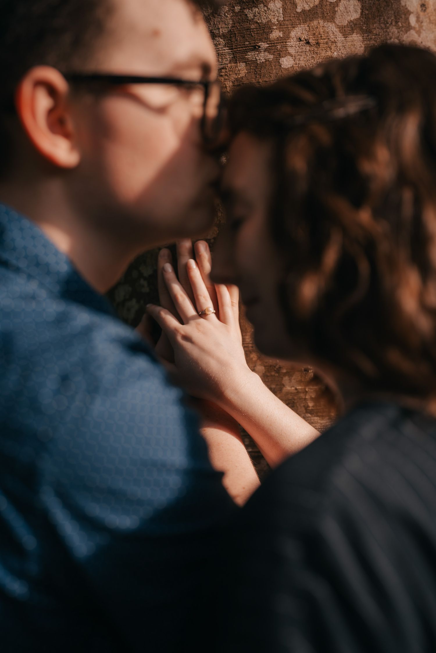 A couple shares an intimate moment in front of a rustic brick wall, with one person wearing a blue shirt.