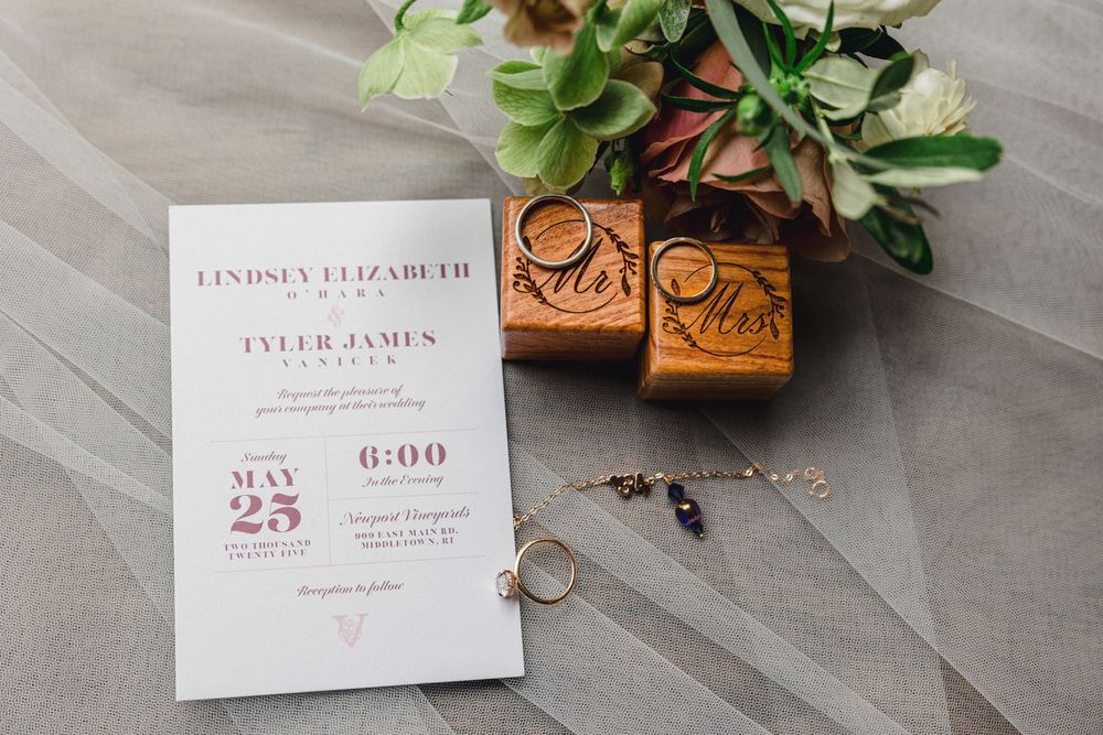 Wedding invitation and rings displayed with delicate pink roses and greenery on a gray surface.