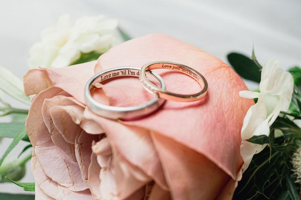 Wedding rings rest on delicate pink rose petals with white floral accents in a romantic bridal detail shot.