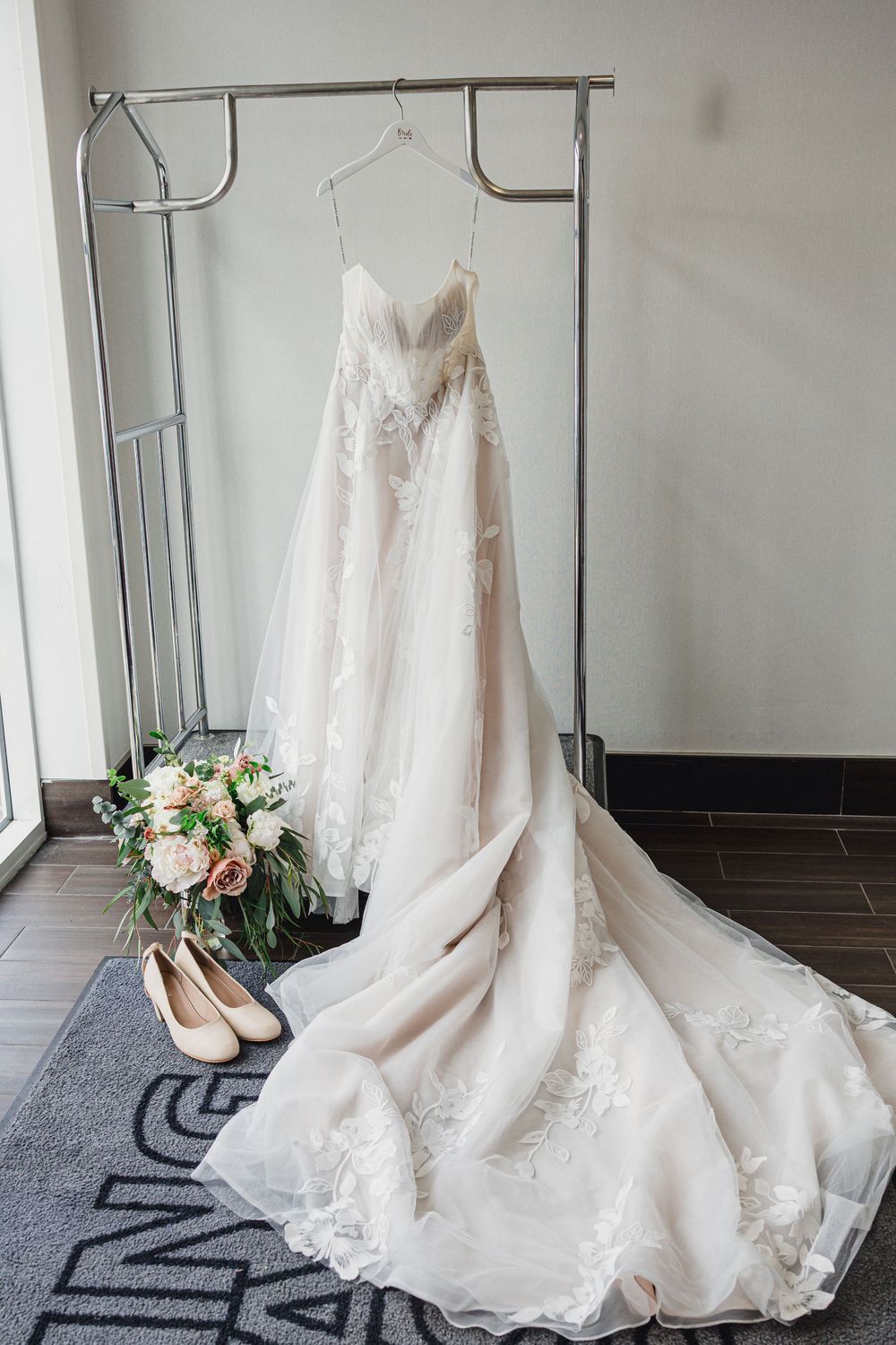 An elegant white lace wedding dress with long train displayed on a hanger with matching shoes and floral bouquet.