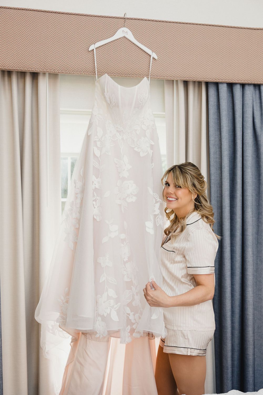 White wedding dress hanging in a bright room with blue and cream curtains.